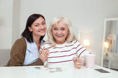  Enthusiastic elder woman and caregiver looking at camera while gathering puzzle